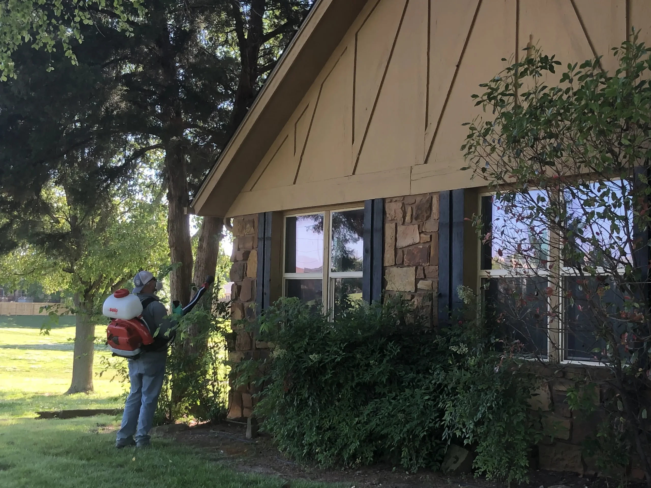 Complete Lawn Care technician treating shrubs beside a brick home with a backpack sprayer