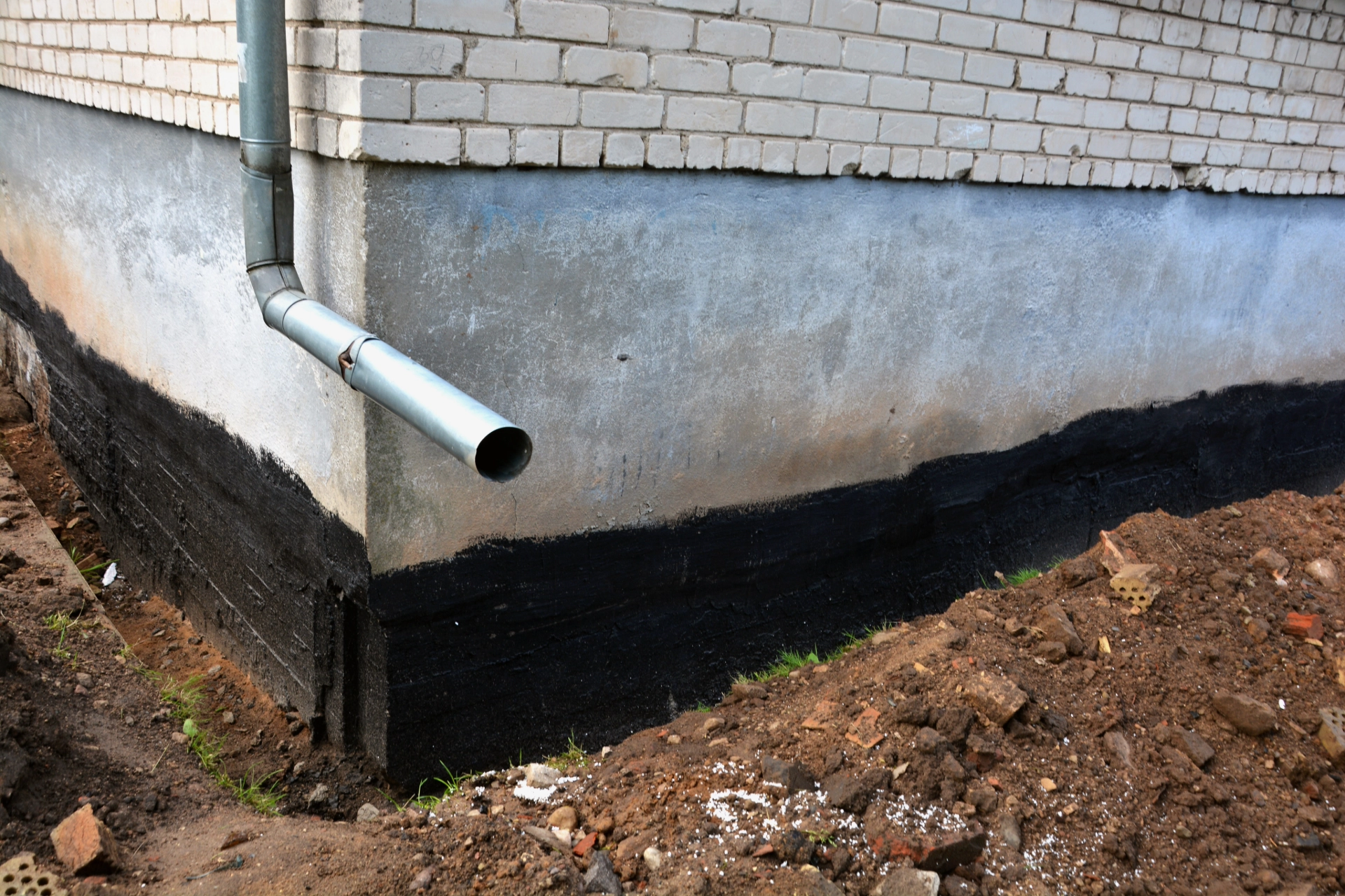 Exposed foundation with black waterproofing coating and a metal downspout drain pipe