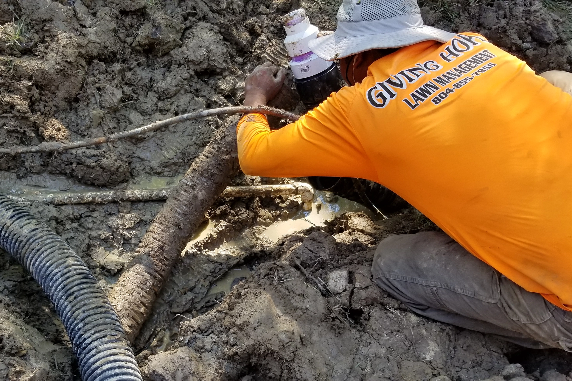 Worker in an orange Giving Hope Lawn Management shirt installing drainage pipe in wet soil