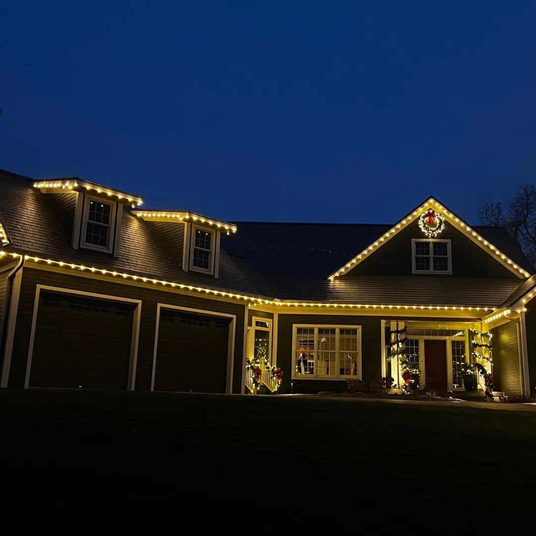 A dark sided single story home at deep blue dusk with warm white lights along every roofline and dormer, a lit wreath with a red bow on the front gable and garland wrapped around the porch posts
