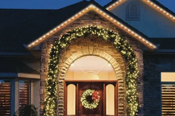 A stone arched entryway wrapped in lit garland with a large wreath on the door and warm white lighting along the rooflines above
