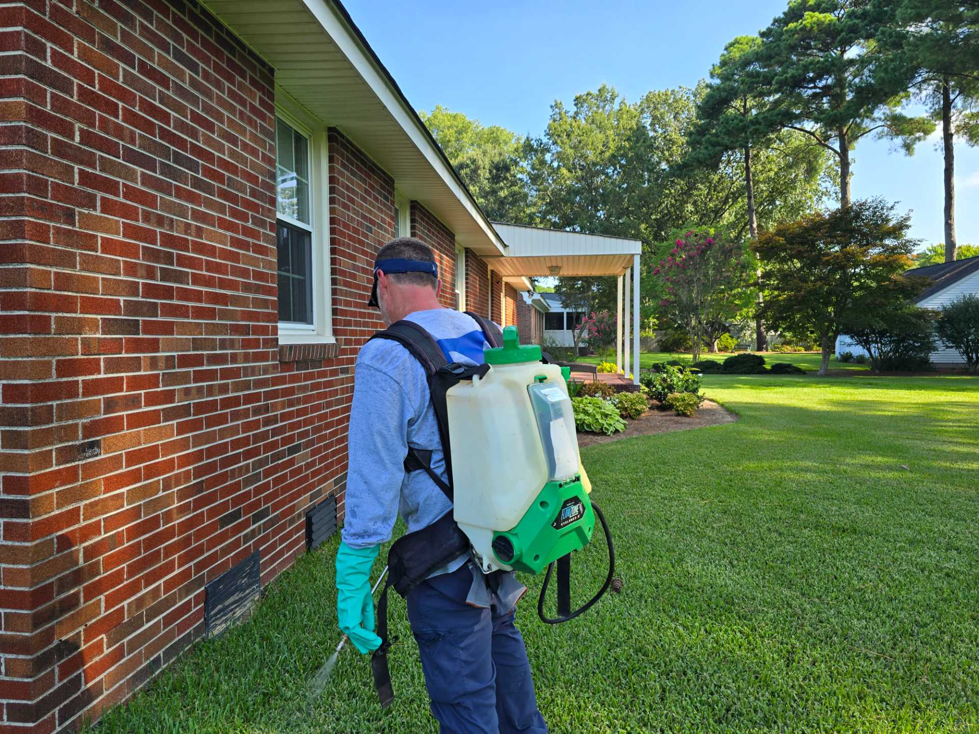Pest control technician applying a perimeter treatment with a backpack sprayer along a brick home foundation