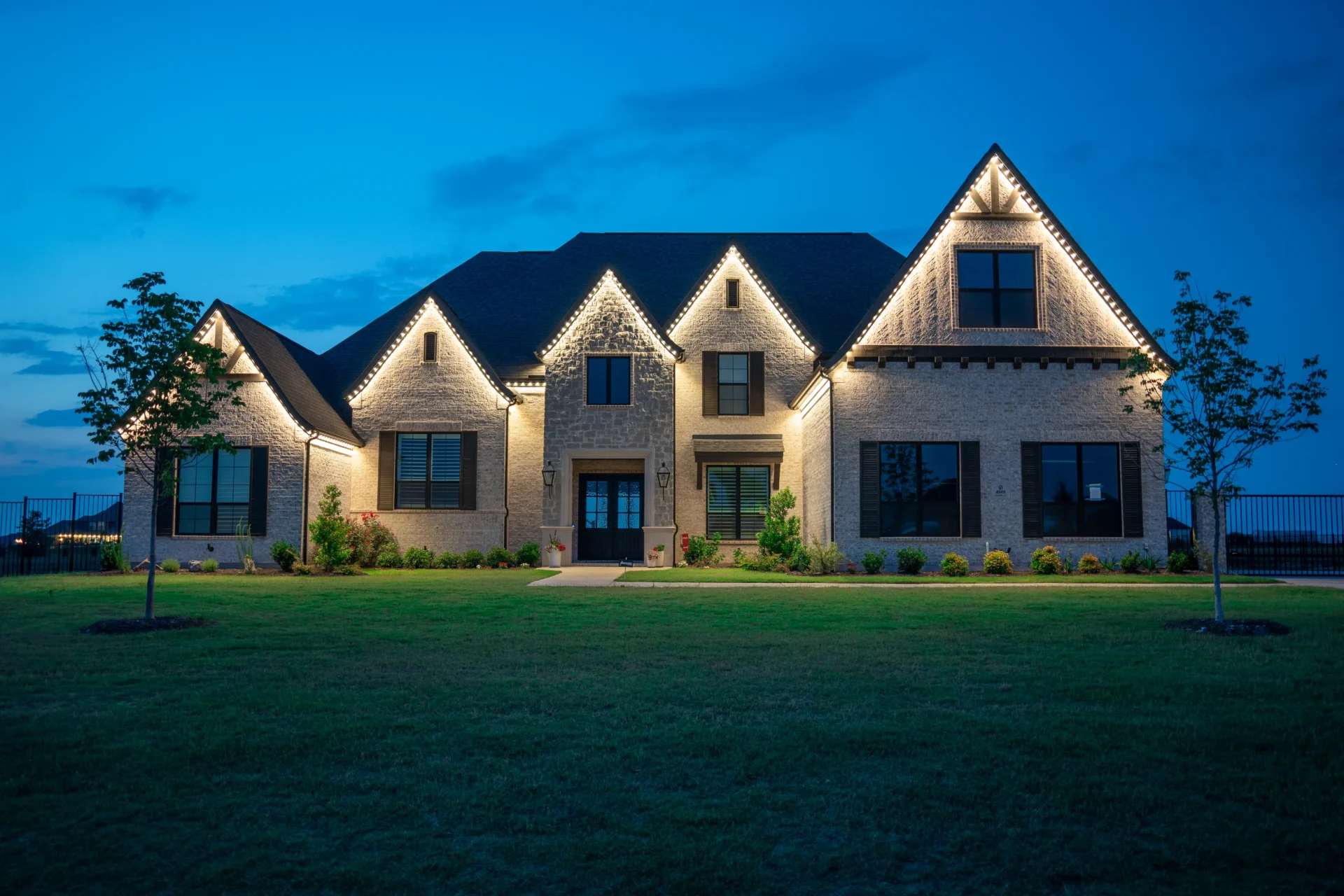 Brick two-story home at dusk with warm white permanent lighting outlining its gables and young trees on the front lawn