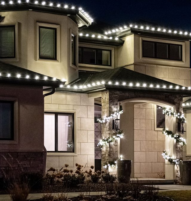 Close-up of commercial-grade C9 LED bulbs clipped to a shingle edge on a New Age Lights seasonal install, warm white against a dusk sky