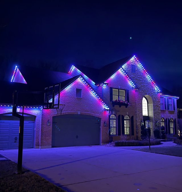 Color-matched permanent lighting track running along the fascia of a two-story brick home in Hoover, AL, lit warm white at dusk