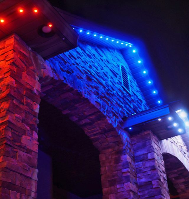 Close-up of a color-matched permanent lighting track seated under the soffit of a brick home in Fort Thomas, Kentucky, with pucks glowing warm white, installed by New Age Lights