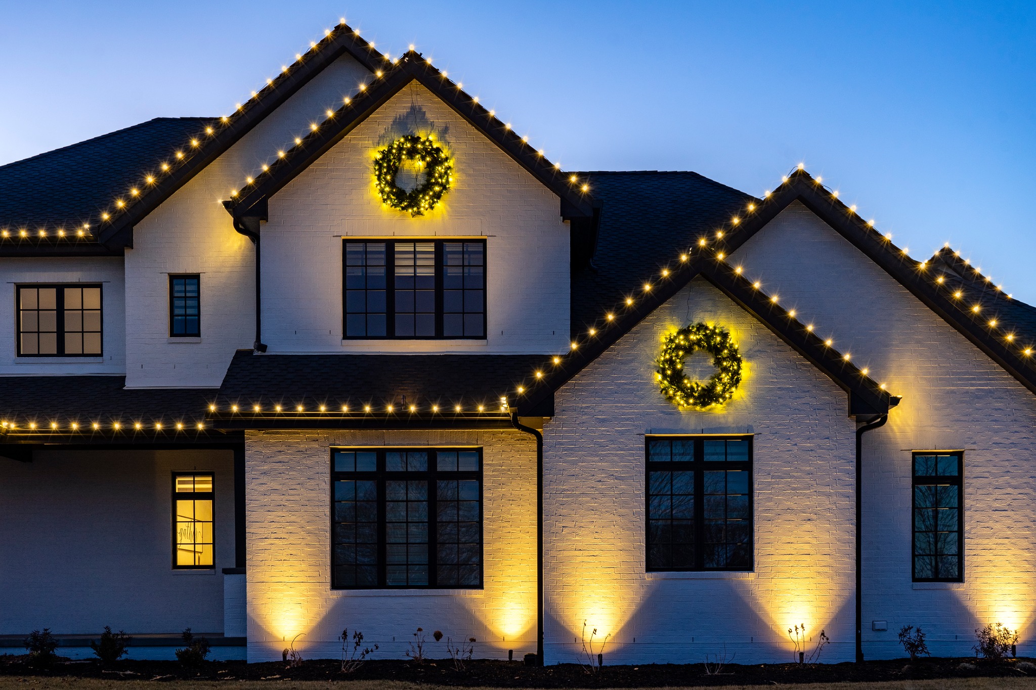 White brick home at dusk with warm white Christmas lights along the rooflines, lit wreaths on the gables and uplighting on the walls