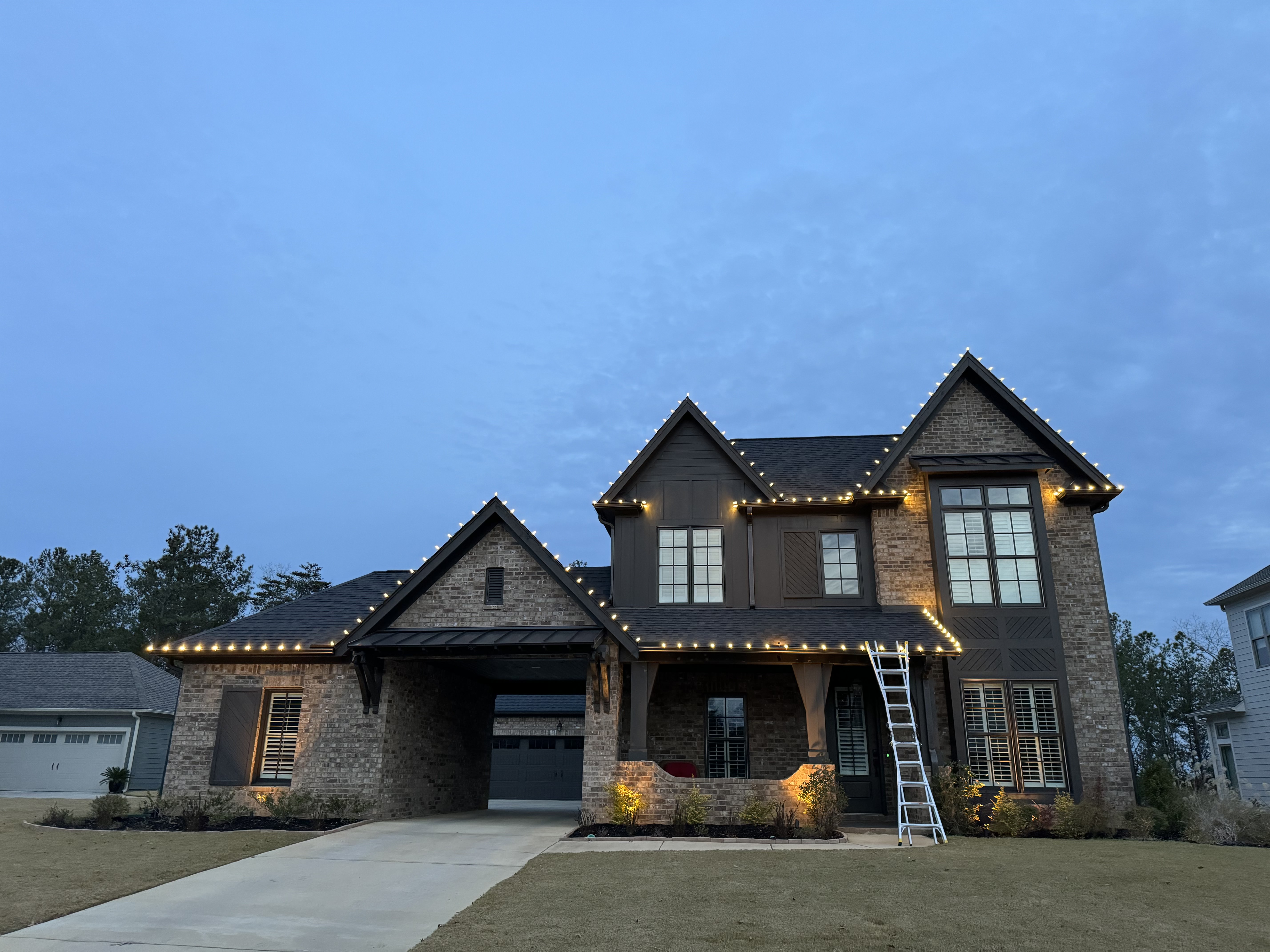 Two-story brick home at dusk with warm white lights outlining the roof peaks and gables, with a ladder at the front porch