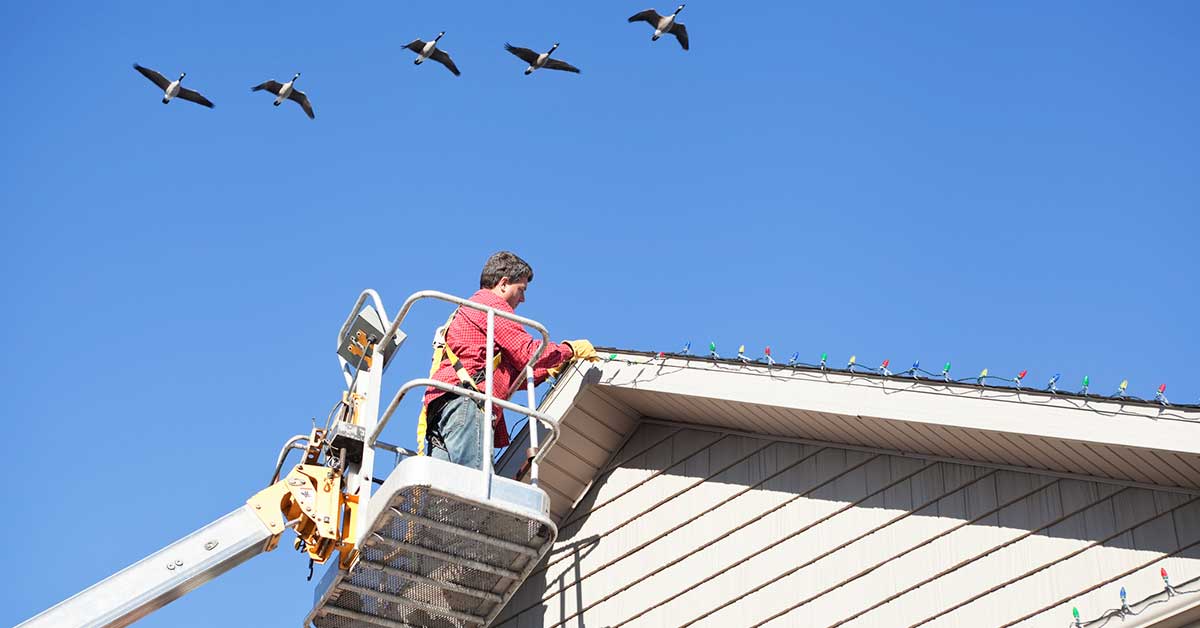 Installer in a bucket lift attaching multicolor Christmas lights along a roofline under a blue sky