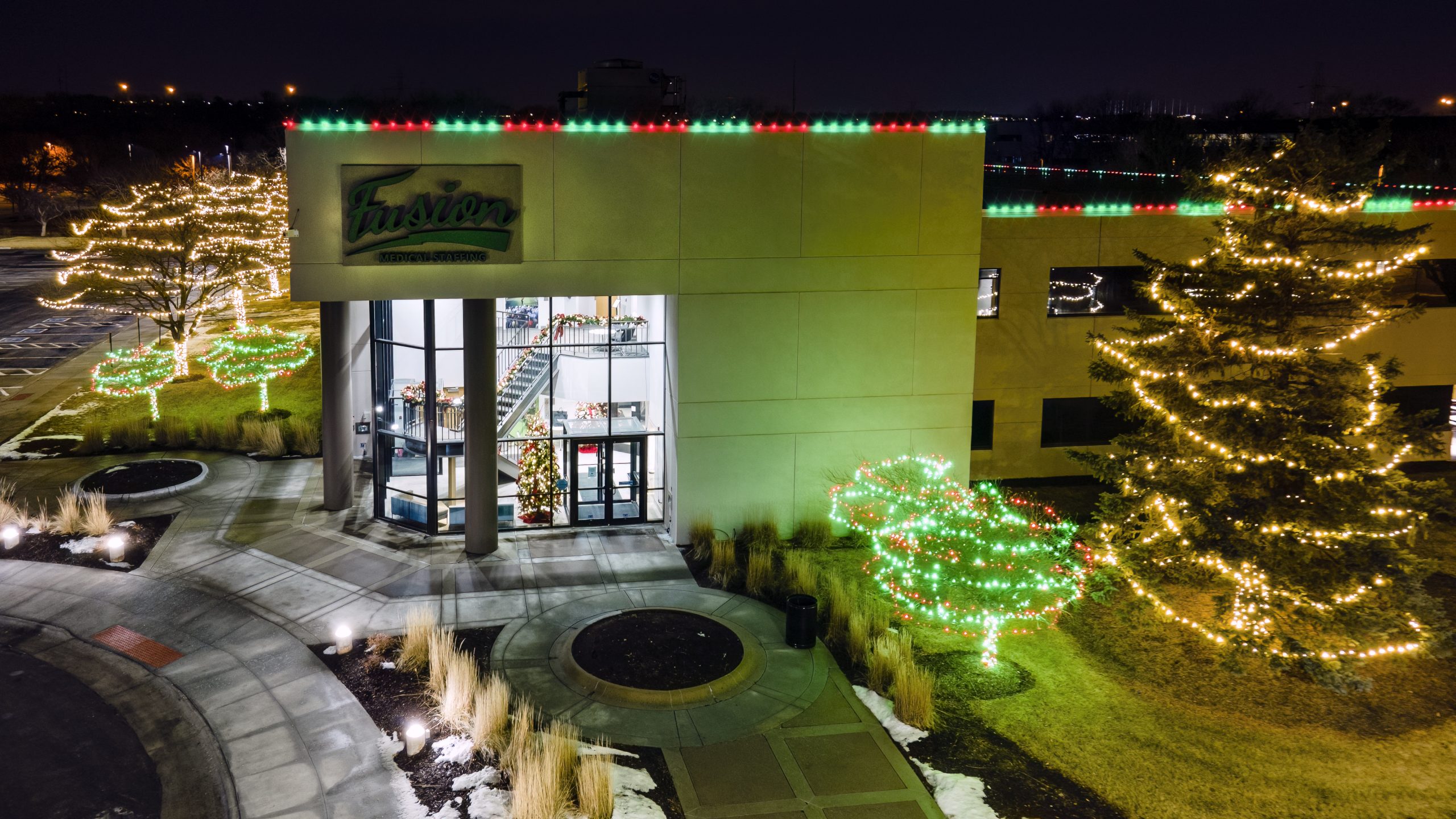 Commercial building entrance lit in green with trees wrapped in lights along the walkway