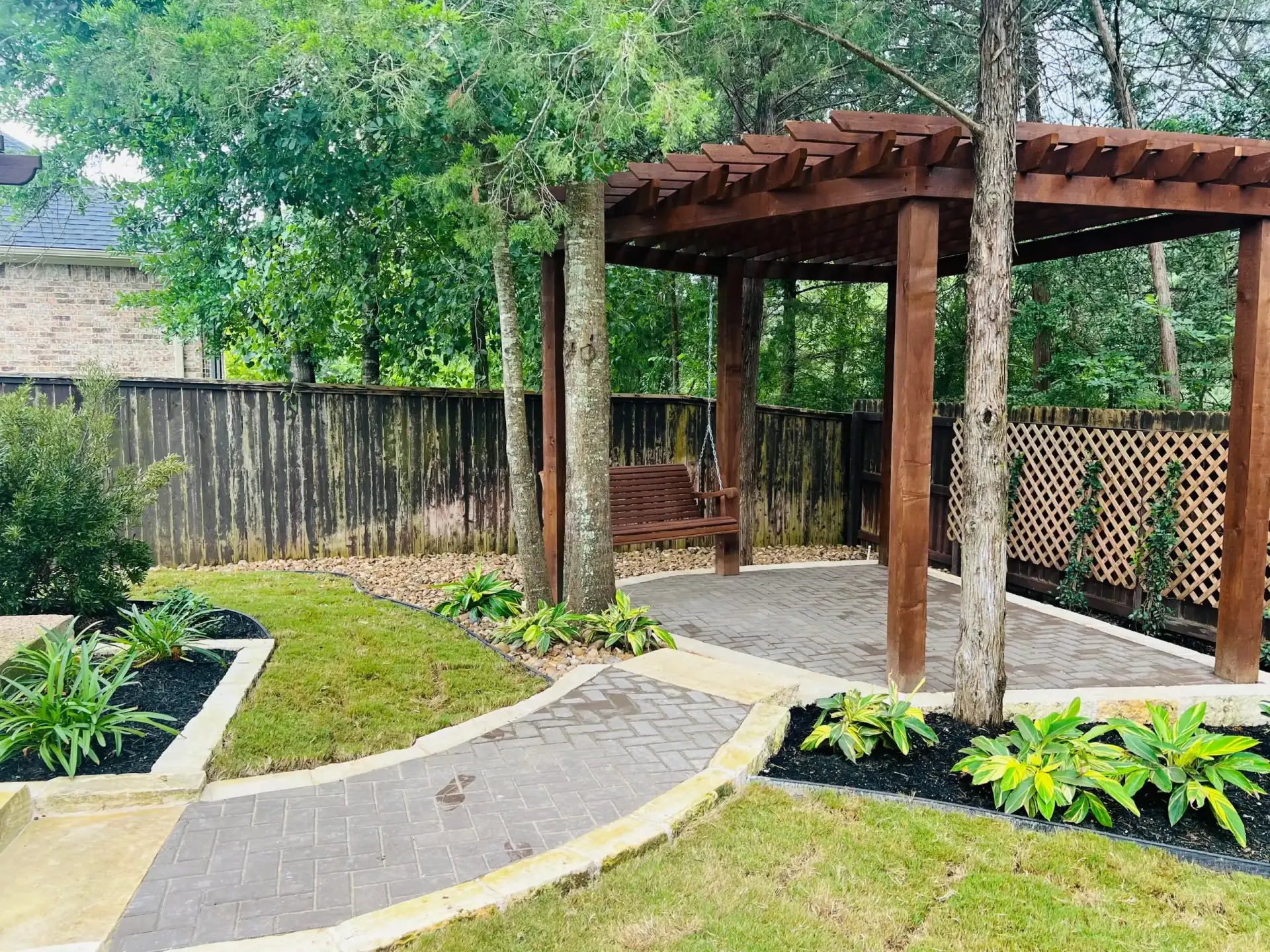 Cedar pergola with a porch swing over a paver patio in a shaded, fenced backyard