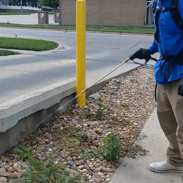 Yard Boss technician spraying a perimeter pest control treatment along a rock bed in Lincoln, NE