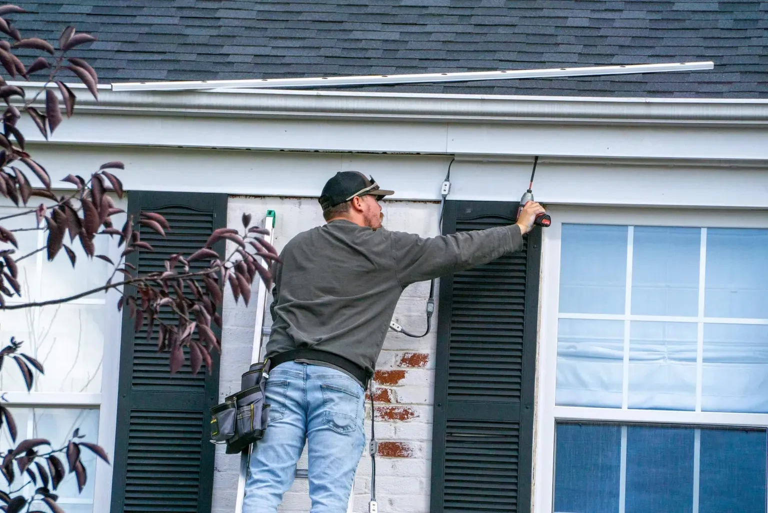 Installer on a ladder fastening permanent lighting track along the fascia of a white brick home with black shutters in Walton, KY