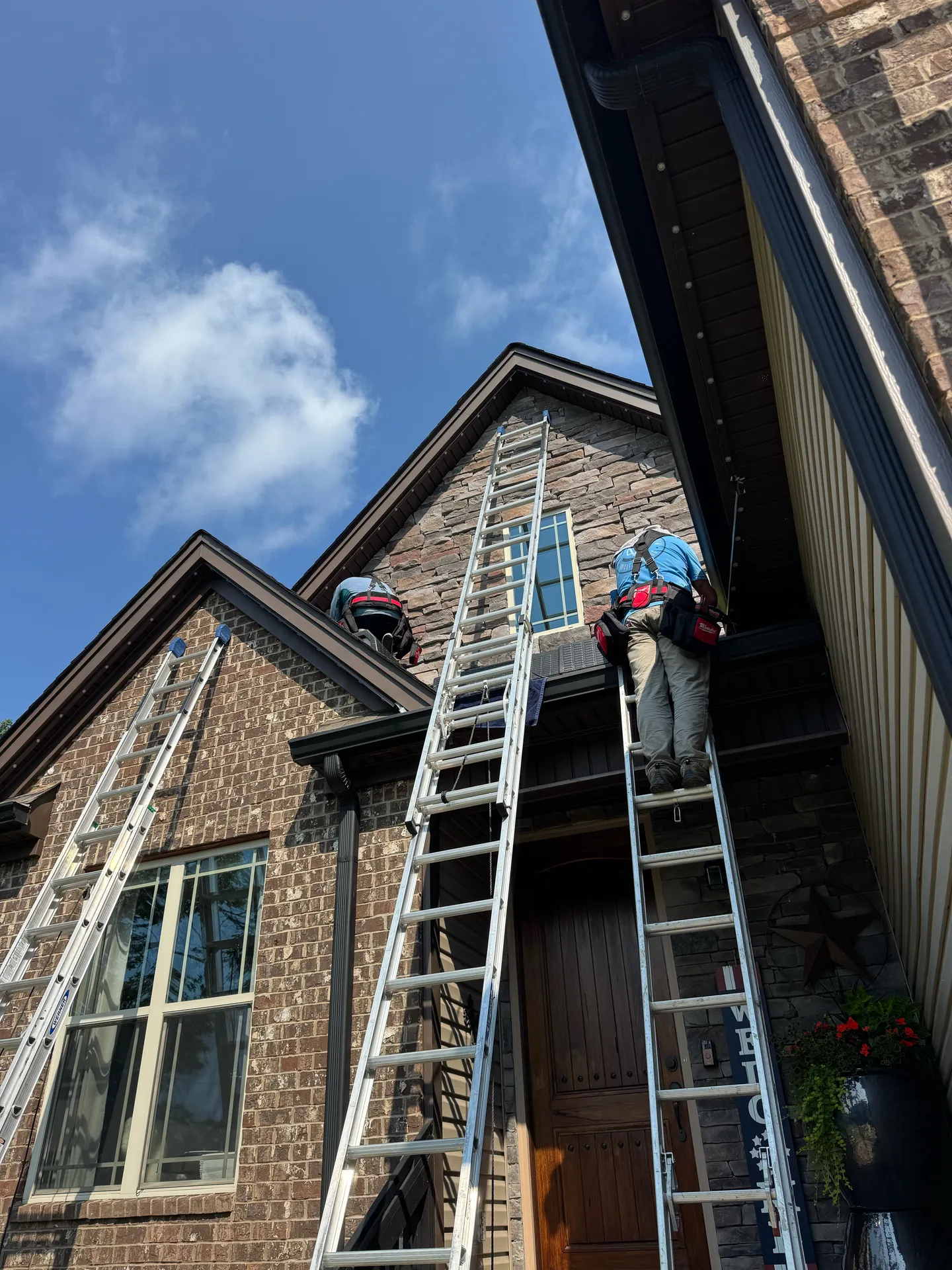 Two installers on extension ladders working along the roofline of a two-story brick and stone home during a JellyFish permanent lighting install in Chattanooga, TN