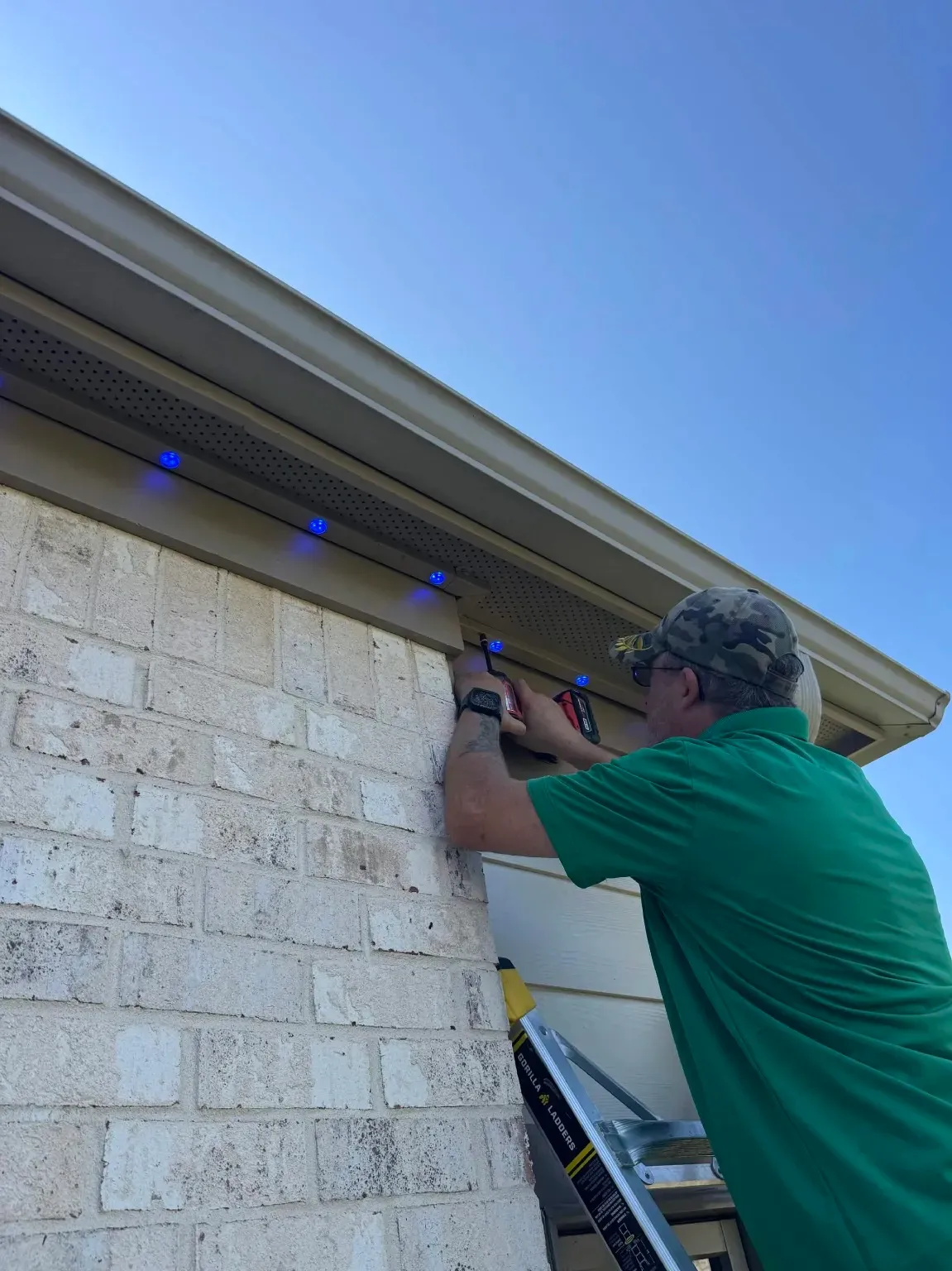 Installer in a green shirt fastening permanent lighting track with blue LED lights under the soffit of a light brick home in Madison, AL
