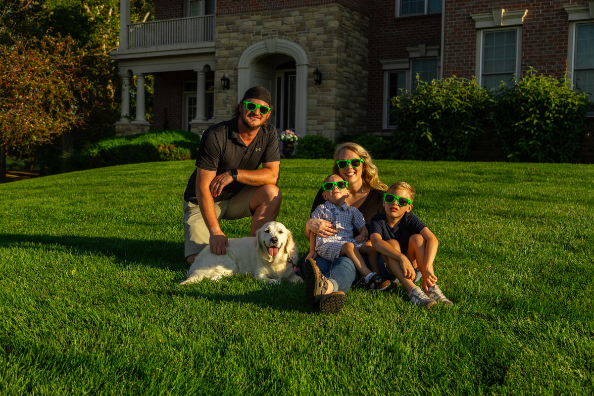 Family in green novelty glasses sitting with their white retriever on a thick front lawn