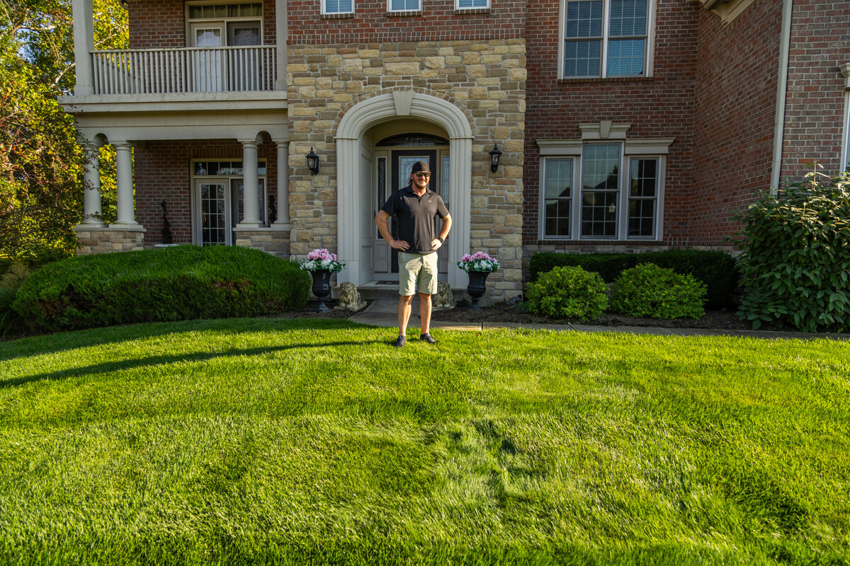 Homeowner standing on a thick, striped front lawn outside a brick home