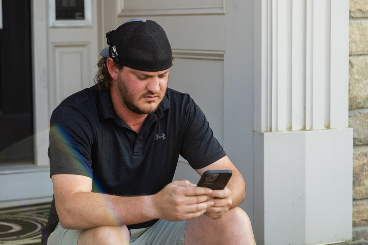Man in a cap sitting on a front porch step, typing on his phone