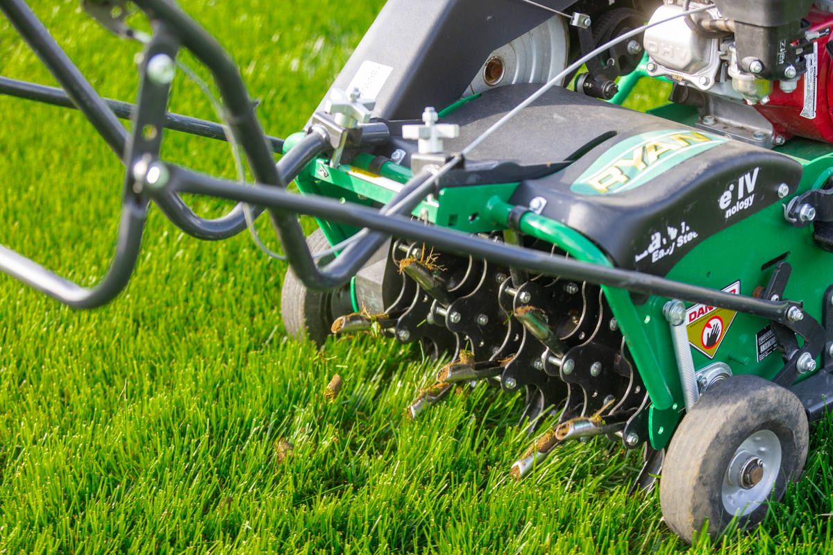 Close-up of a walk-behind core aerator's tines pulling soil plugs from a lawn