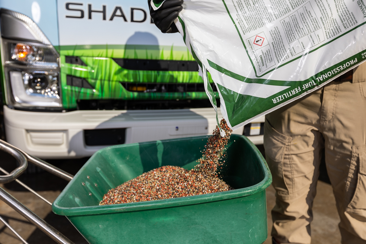 Granular fertilizer pouring from a bag into a walk-behind spreader, Shades of Green truck behind