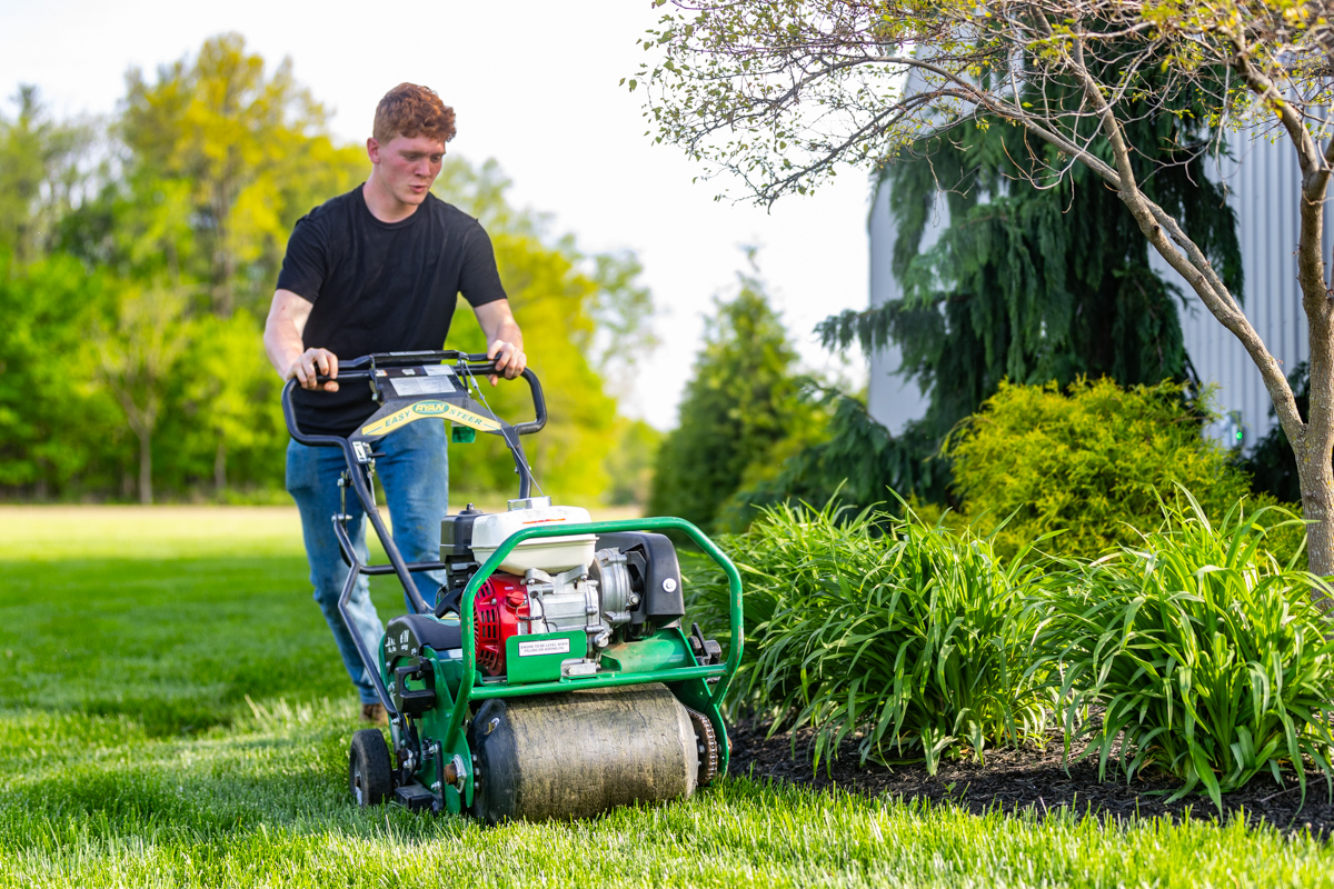 Young homeowner pushing a walk-behind core aerator past a landscape bed