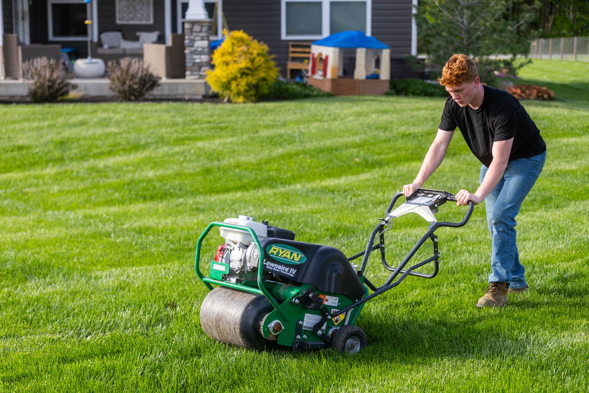 Young homeowner pushing a walk-behind core aerator across a striped backyard lawn