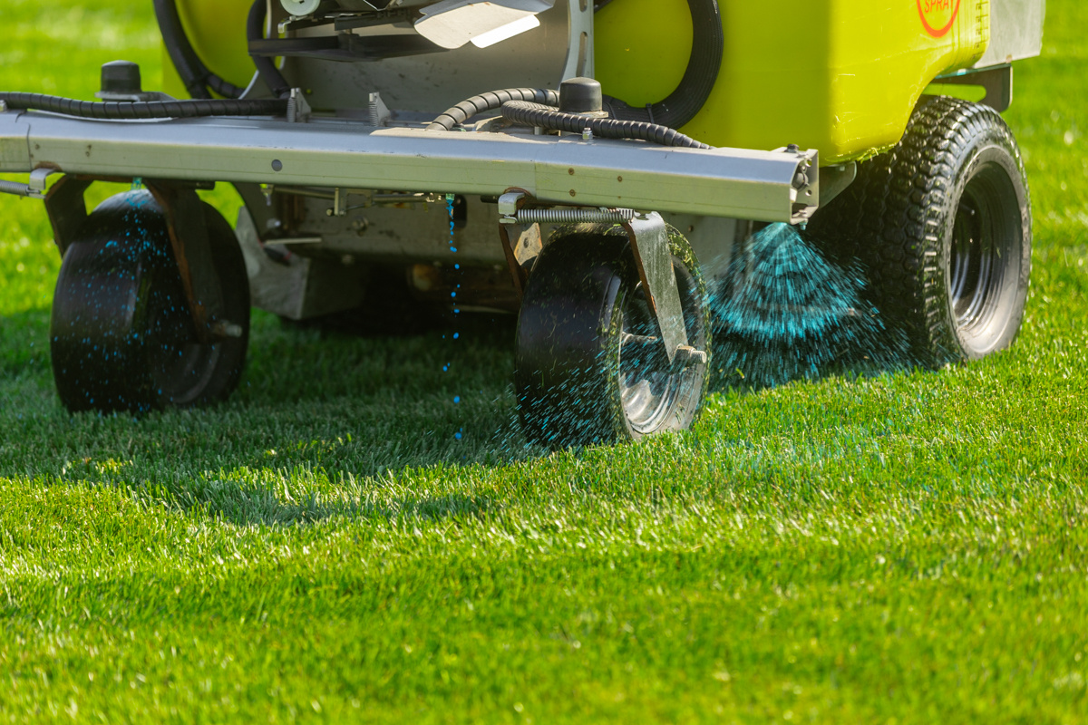 Close-up of a ride-on sprayer's nozzles misting blue-dyed liquid onto green grass