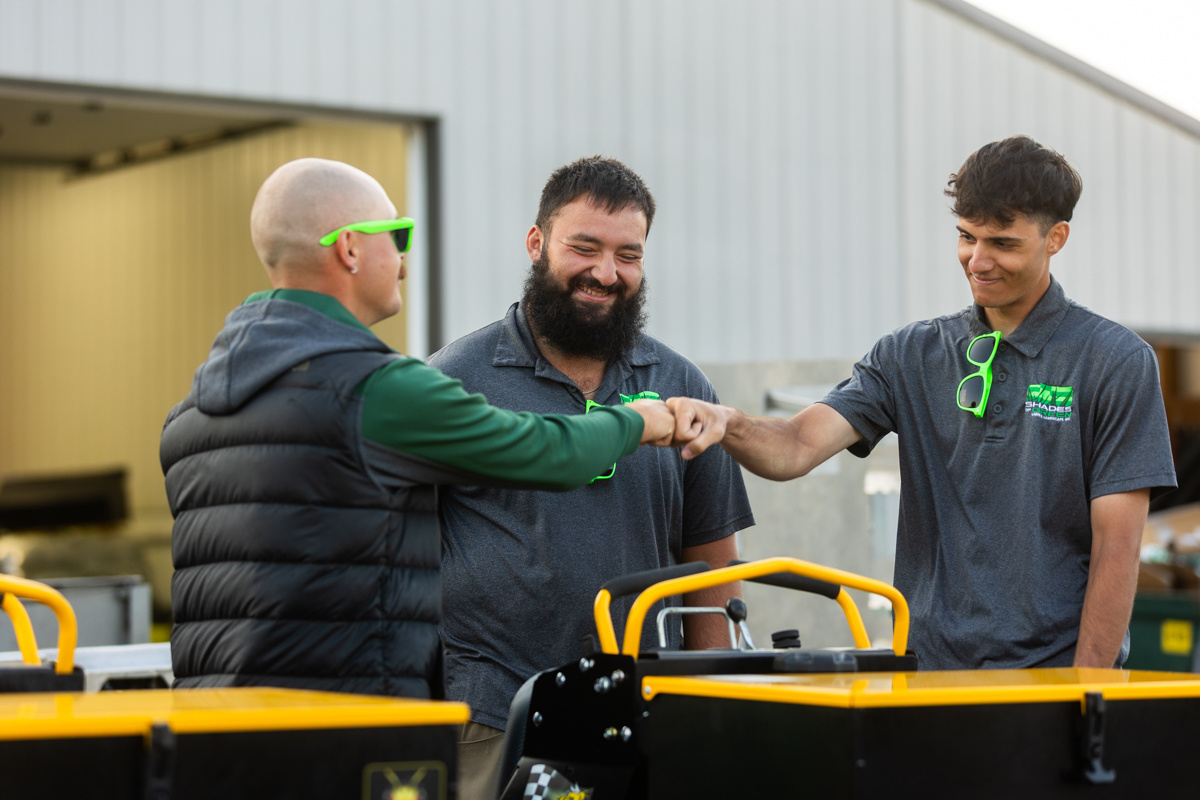 Three Shades of Green team members bumping fists beside lawn equipment outside the shop
