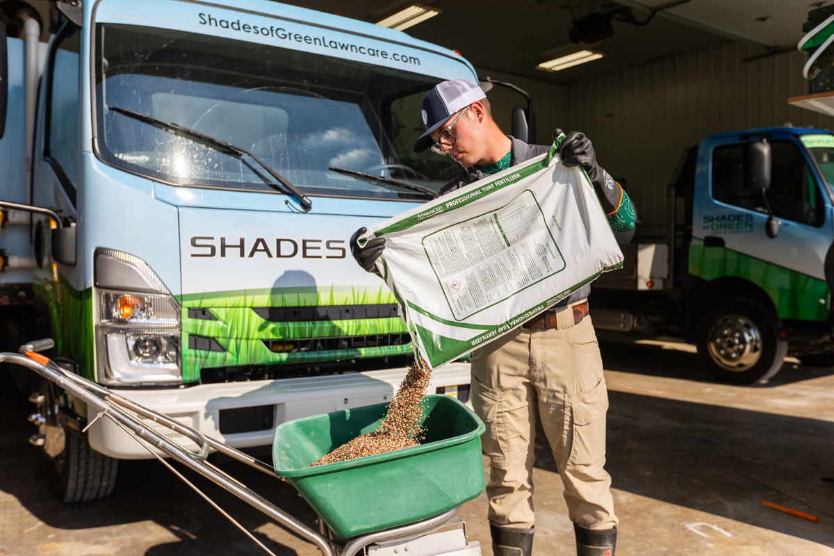 Technician pouring granular fertilizer into a walk-behind spreader in front of a Shades of Green truck