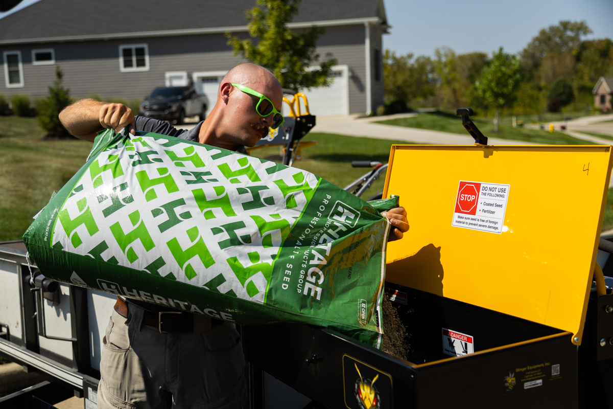 Technician emptying a green bag of grass seed into a yellow-lidded spreader hopper