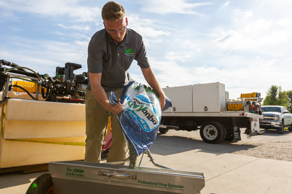 Technician pouring a bag of grass seed into a drop spreader beside the service truck