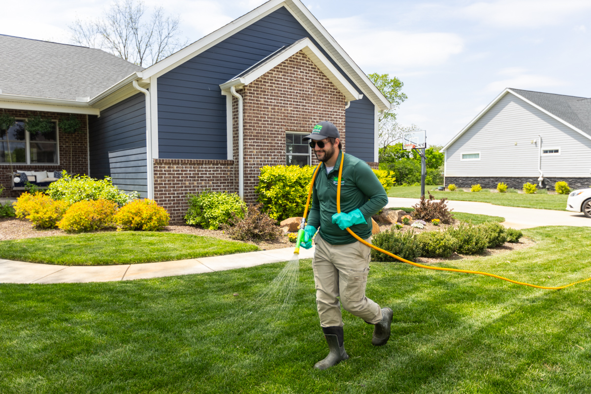 Technician spraying a liquid lawn treatment with a hose in front of a blue home