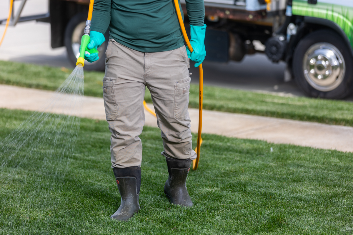 Technician in rubber boots spraying a lawn, service truck parked behind