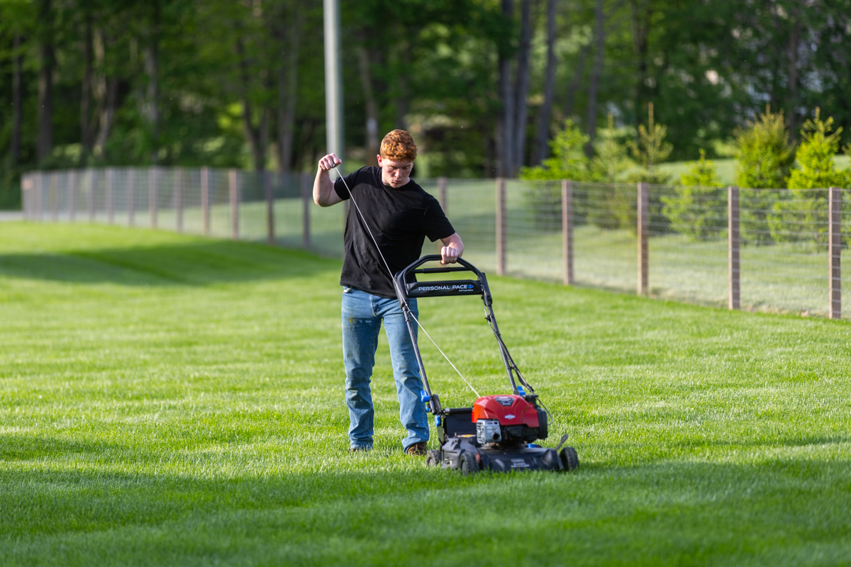 Homeowner pull-starting a push mower on a fenced backyard lawn