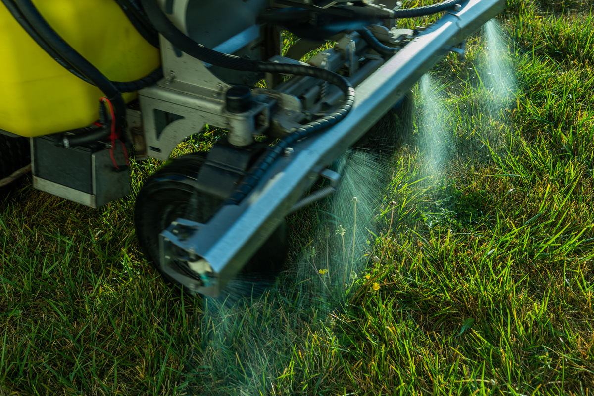 Ride-on sprayer boom misting a liquid treatment onto a lawn, seen from ground level