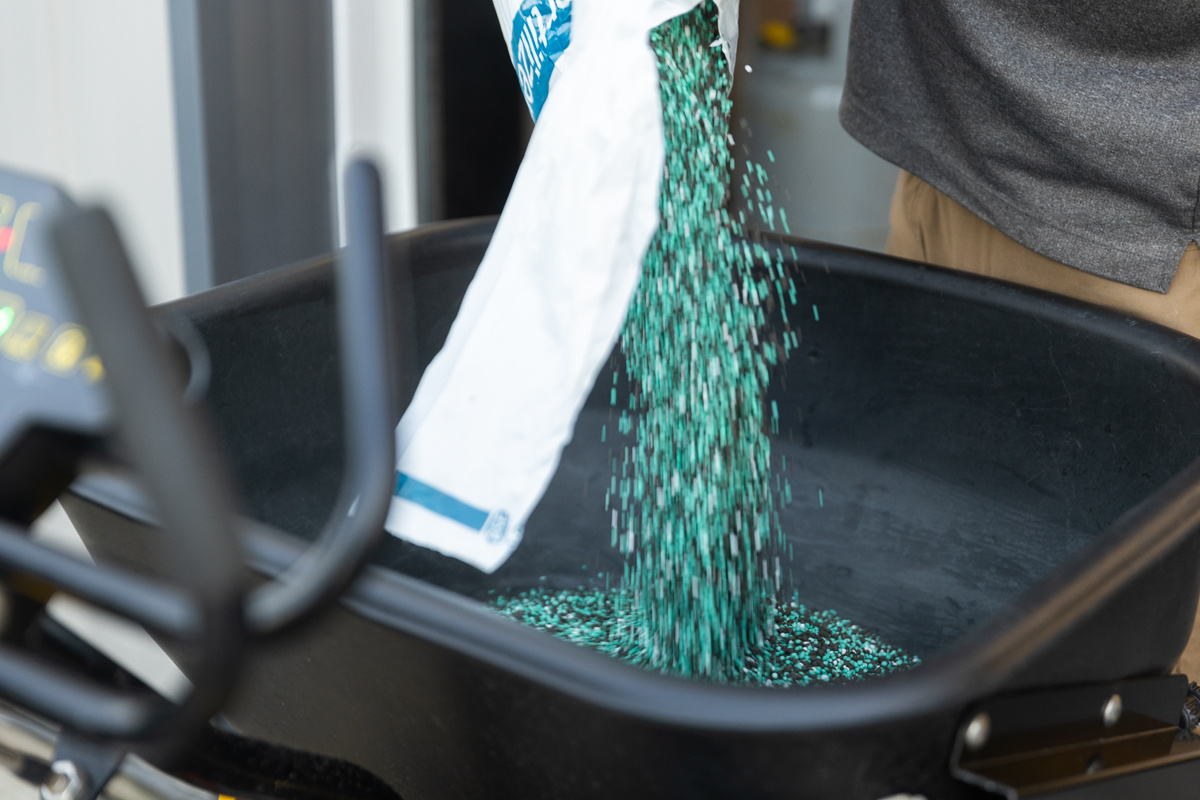 Blue-green granular fertilizer pouring from a bag into a spreader hopper