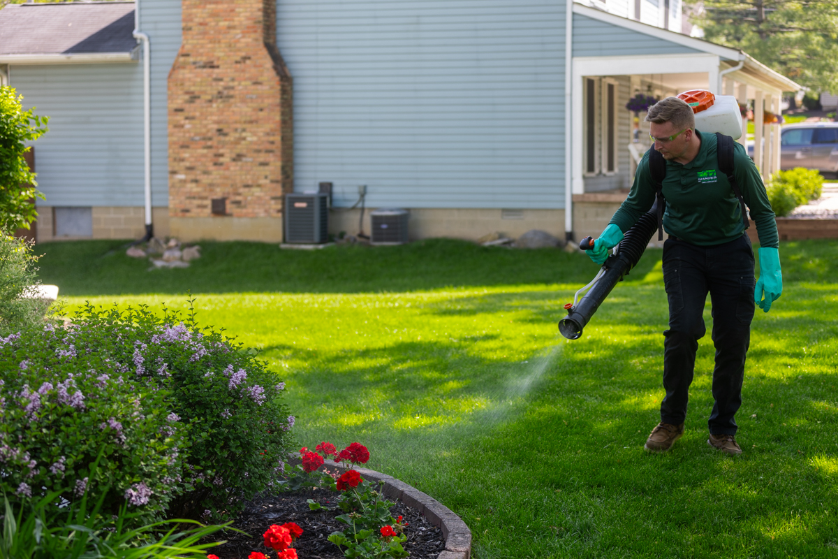 Technician with a backpack mist blower spraying along a backyard flower bed