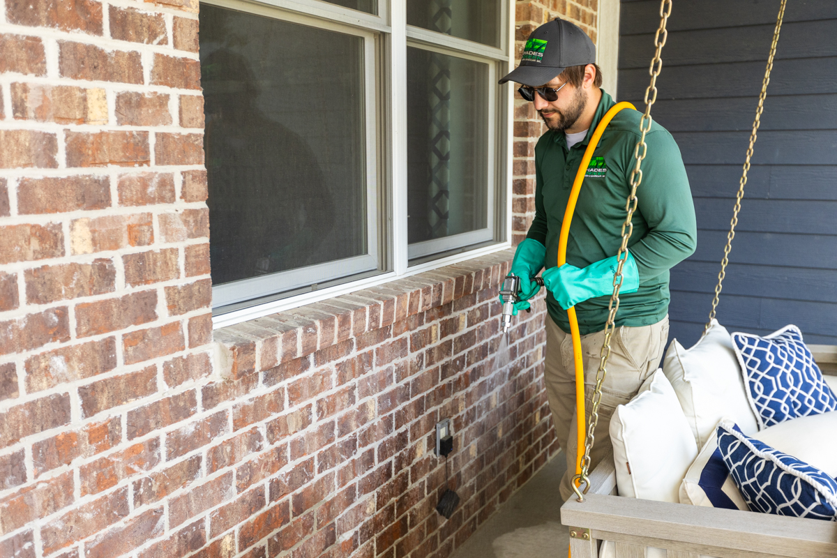Technician spraying a perimeter treatment along a brick wall beside a porch swing