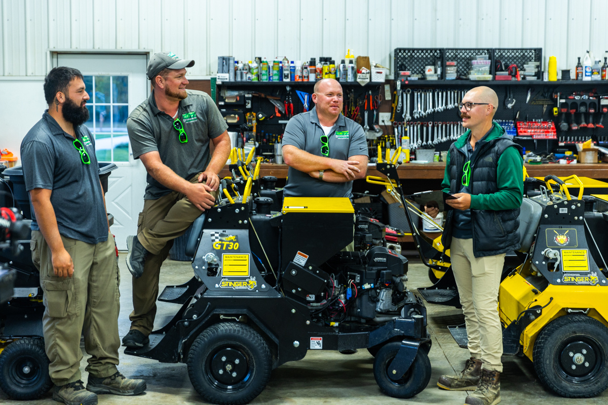 Four crew members talking among ride-on spreaders and sprayers in the shop