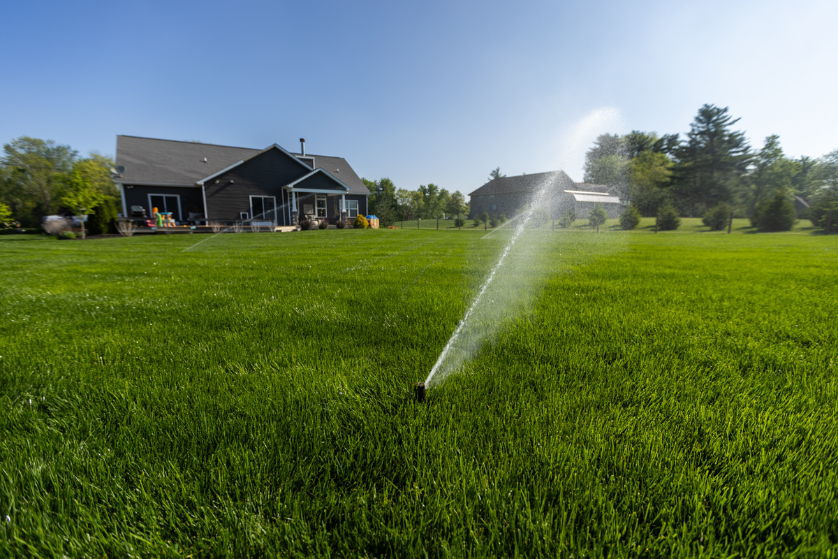 Pop-up sprinkler watering a wide lawn in front of a dark-sided home