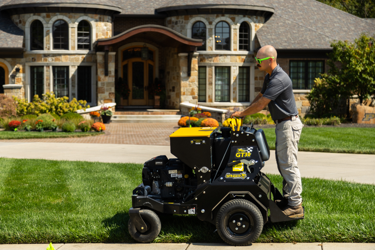 Technician driving a stand-on core aerator across the front lawn of a stone home
