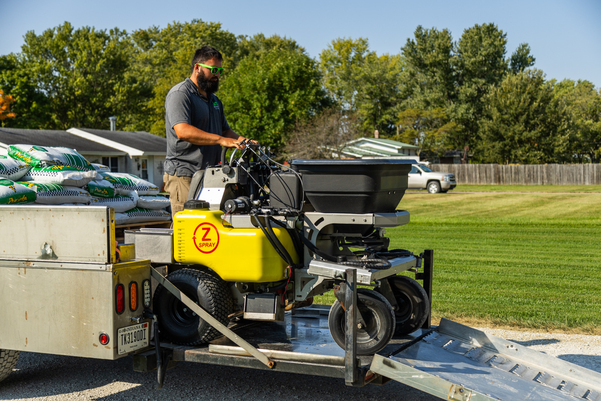 Technician backing a ride-on spreader-sprayer off a trailer loaded with product bags