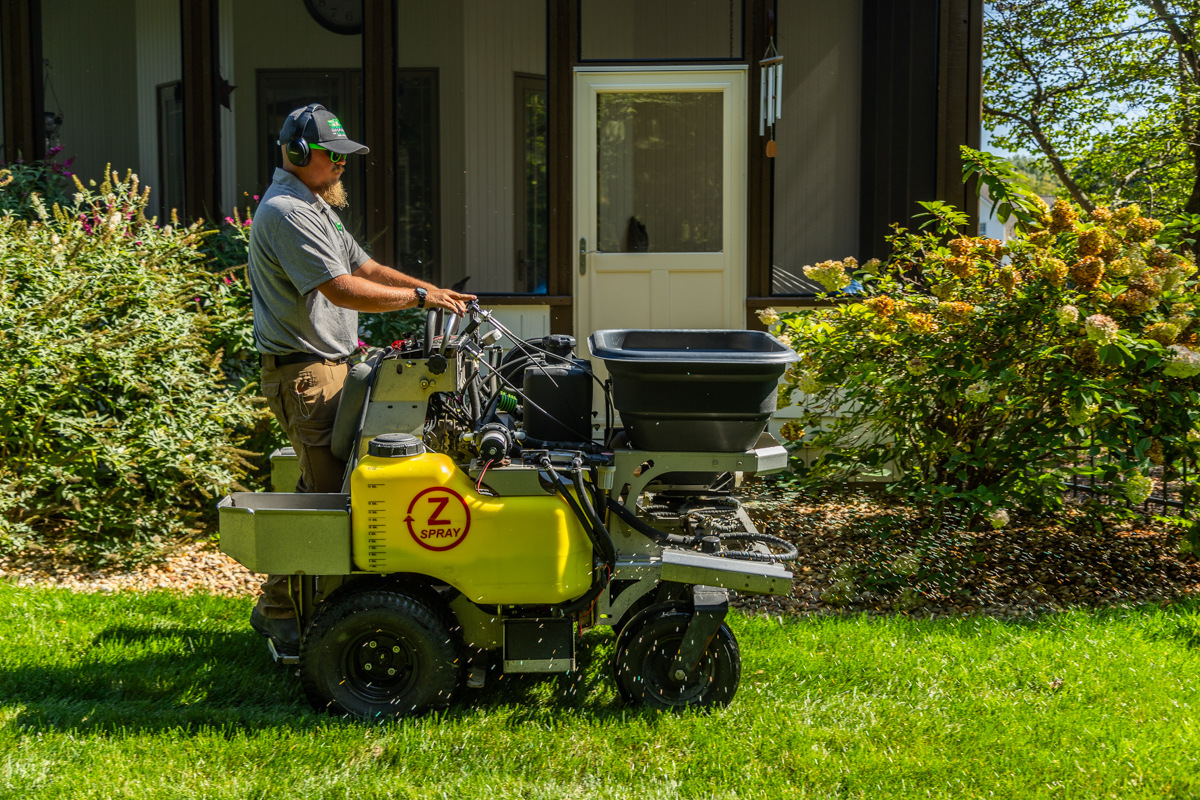Technician on a ride-on spreader by a backyard patio and flowering shrubs