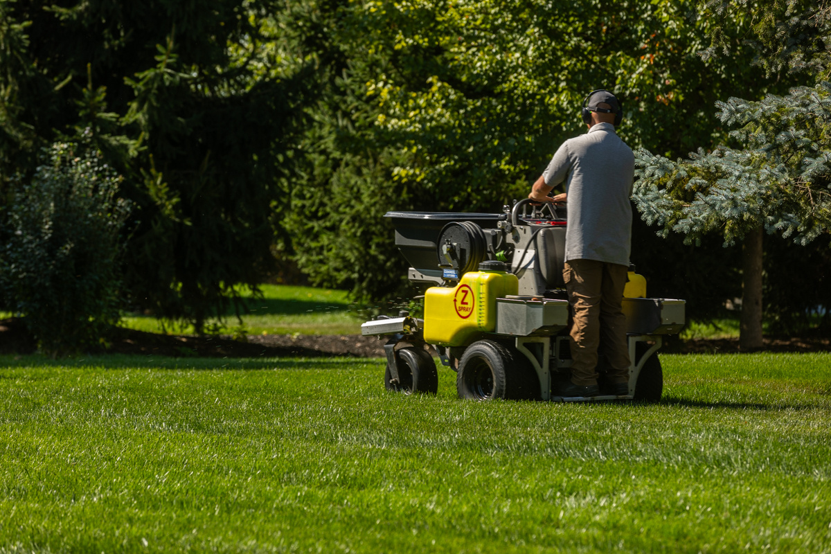 Technician on a ride-on spreader crossing a large, tree-lined lawn