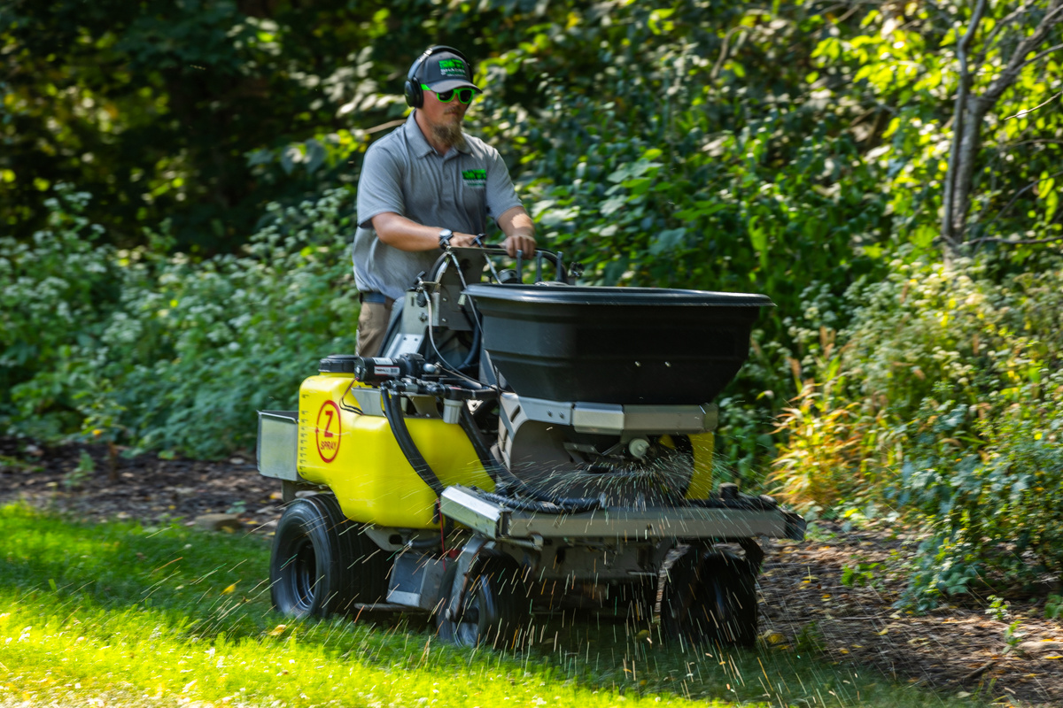Ride-on spreader throwing granular fertilizer along a landscaped bed