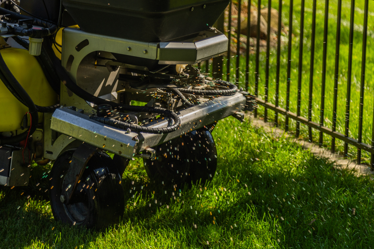 Close-up of a spreader broadcasting fertilizer granules beside an iron fence