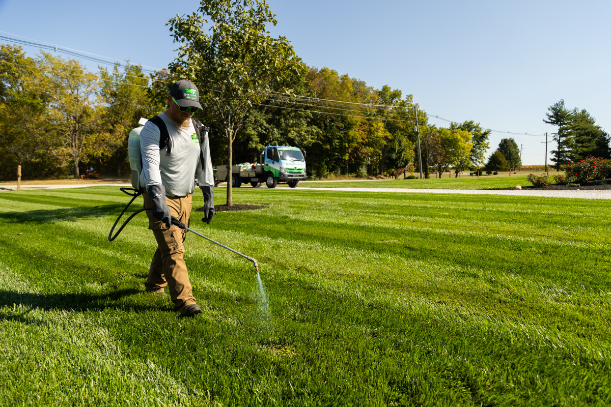 Technician with a backpack sprayer spot-treating a striped lawn, service truck behind