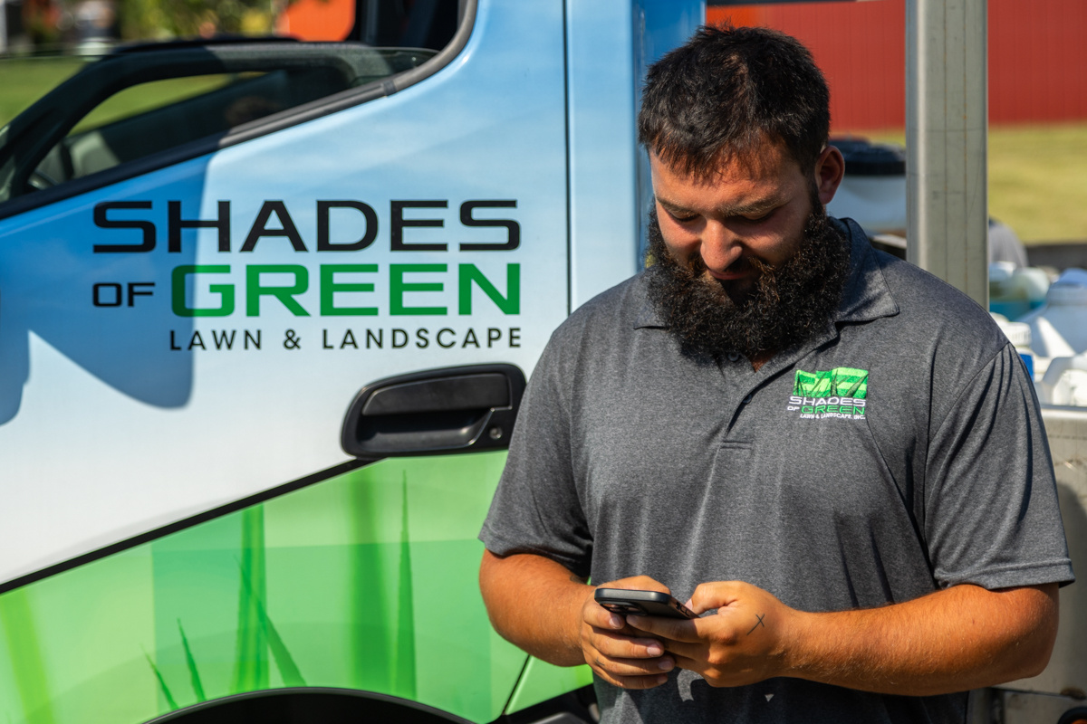 Technician checking a phone beside a Shades of Green Lawn & Landscape truck