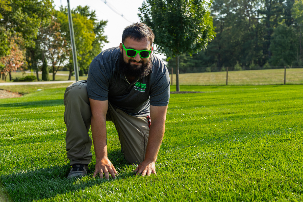 Technician in green sunglasses kneeling with hands on a thick, striped lawn