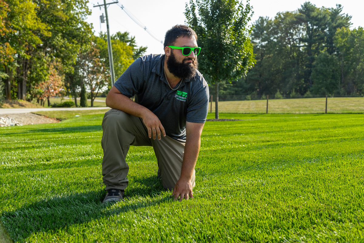 Technician in green sunglasses kneeling on a thick, striped lawn, looking across it